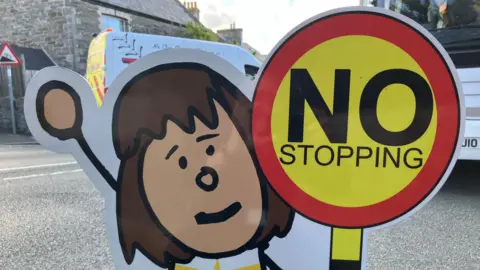 A Parking Buddy sign. It is a cartoon child with brown hair holding a yellow and red 'no stopping' sign. The sign is on the road outside a school and a white bus and white van are behind the sign.