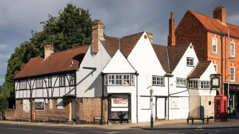 Historic England The Old Ship Inn, a whitewashed, half timbered structure with a sloping tiled roof sat on the junction of two roads