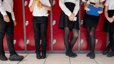Four pupils stand against red lockers at a school. Three girls and boy are stood in a line, one is holding paperwork and another a phone. Their faces are not visible.