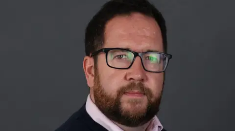 Scottish Borders Council A dark-haired man with glasses, wearing a black top and pink shirt, looks at the camera