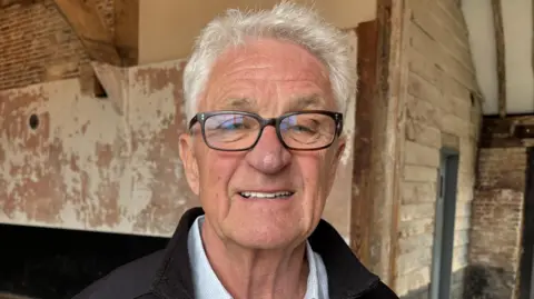 Luke Deal/BBC A man with short grey hair smiles at the camera. He wears glasses, a black jumper with a white shirt underneath. He is standing in a barn.