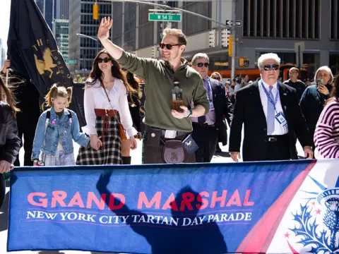 Getty Images A man in a kilt and green shirt, holding a bottle of whisky waves at onlookers as he leads a march