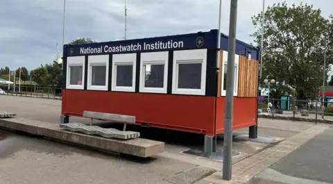NCI Minehead The exterior of the station is pictured. It is a small one-room building. It is painted navy blue and burgundy.