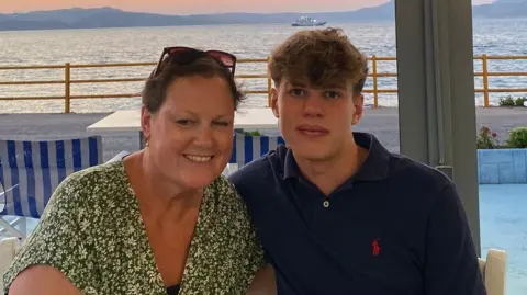 Finn is on the right of the shot, wearing a blue polo shirt and looking at the camera with a neutral expression. He has his arm behind his mum, to the left, who is wearing a green floral top, and is smiling at the camera. They appear to be on holiday, backed by the sea.