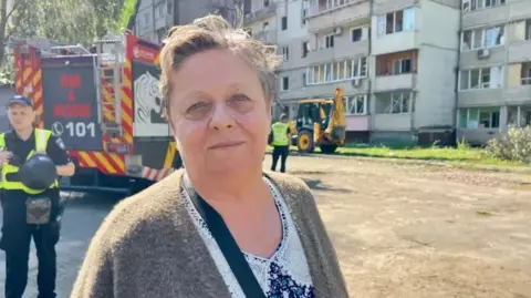 Valentina, uma senhora com cabelos curtos em um cardigã marrom e camisa floral estava em frente ao prédio com bombeiros e um caminhão ao fundo