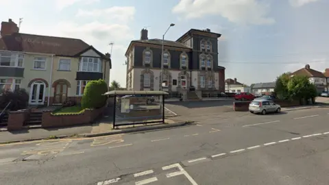 A bus shelter in the middle of a main road. Large buildings can be seen lining the street, as well as road markings.