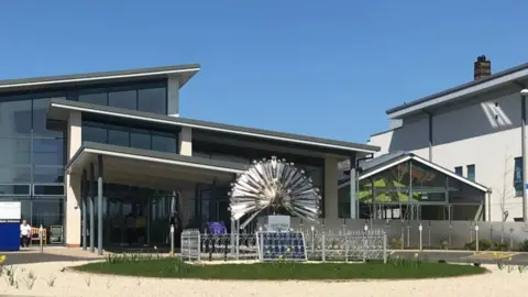 The front of a large, glass-fronted building with a large metallic ball-shaped sculpture in front of it. The roofs are black and flat and there is a gravel and grass area in front.