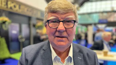 Paul Leeman, chair of the Northern Ireland Conservatives, at the Tory conference in Manchester. Mr Leeman has grey/fair hair, white eyebrows and is wearing black-rimmed glasses. He is wearing a grey suit jacket and open-necked white shirt 