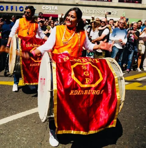 Drummers from the Scottish Association of Marathi Regional Art and Traditions (SAMRAT) wearing bright orange tops perform in a street parade, carrying large bass drums draped with red banners reading “Samrat SGF Edinburgh.” Spectators stand along the route watching the performance.