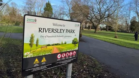 A large sign is at the entrance to a public path. In the background is a concrete path which forks off left and right. Surrounding it is green grass and large trees with no leaves on them. A person is on the right, standing on the grass in a long black coat. The sign has a photo of a lush green field with a bench on it and birds flying in the sky. Text reads "Welcome to Riversley Park"