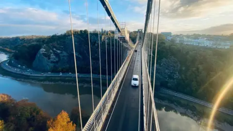 Nicky Willets/Taziker A view of the Clifton Suspension Bridge as taken from high up on the wire suspension cables. It shows the road along the bridge, with a white car driving towards the camera. The sun is setting on the right of the camera and casting a light streak across the corner. 