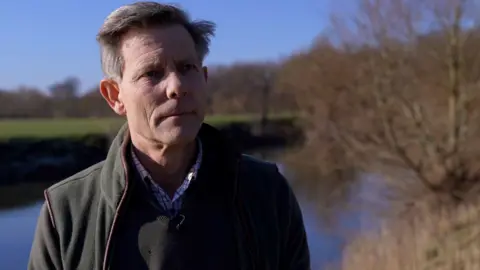 BBC A headshot of a man standing in front of a body of water, a lake or river. He has short, dark grey hair and is looking into the camera. He is wearing a check shirt with a jumper and fleece bodywarmer over the top. The sky is blue in the background and the bank of the water can be seen on other side of the water.