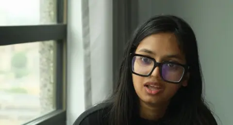 A young woman with long black hair wearing black glasses is next to a window.
