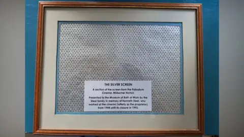 Duncan Steel A wooden picture frame with a section of silver screen with a note explaining that it came from the Palladium and was presented to the Museum of Bath at Work in memory of Ken Steel who worked there from 1944 to 1993