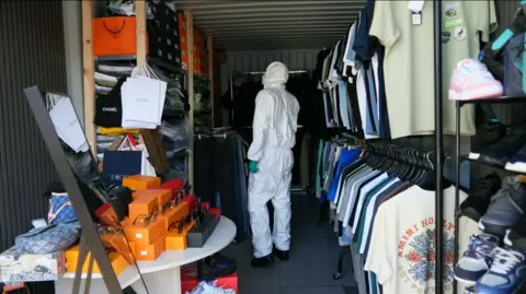 Rows and rails of imitation designer items including trainers, sunglasses, store branded carrier bags and rails of clothes. There is a full length mirror in shot and a man wearing a white suit and disposable gloves stands inside the container.