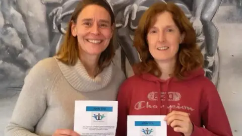 Two women, one in a red hoodie and one in a cream sweater, pose together smiling and holding up a white certificate for the camera