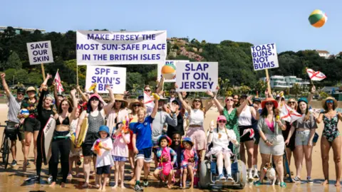 Government of Jersey/ Macmillan Jersey A large group of adults and children cheer on a beach holding signs which read 'slap it on, Jersey' and 'save our skin'.
