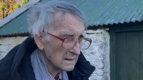 Mr McGoldrick is an elderly man who has short grey hair and slightly wrinkled skin. He is pictured leaning over a grey metal gate. He is wearing red glasses, striped shirt, grey jumper and black coat. Behind him is a white brick shed with a green door and roof.