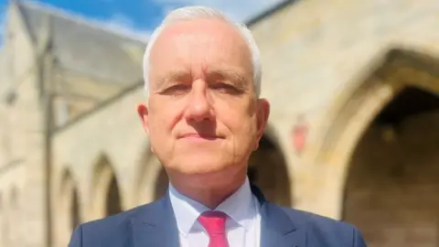 Prof Peter Edwards, a man with white hair and wearing a blue suit jacket, light shirt and red/pink tie, in a university setting.
