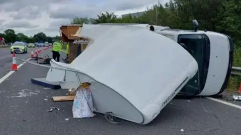 A crashed motorhome on its side at the edge of the road, with police around it. The motorhome is badly smashed up and cars can be seen queueing behind it. 