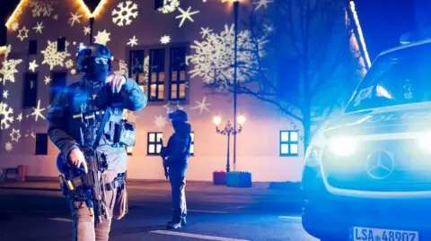 Reuters Armed officer stands in front of building with Christmas decorations projected onto it and an ambulance to the side in Magdeburg