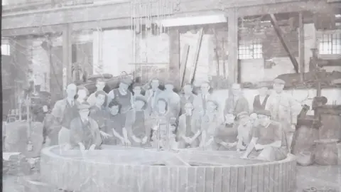 South West Heritage Trust A photographic image shows 23 workers dressed in jackets and caps posing for photographer Henry Wykes at the Bodley Foundry on Commercial Road, Exeter. They are standing and kneeling in front of a huge wheel which is placed on the ground.

