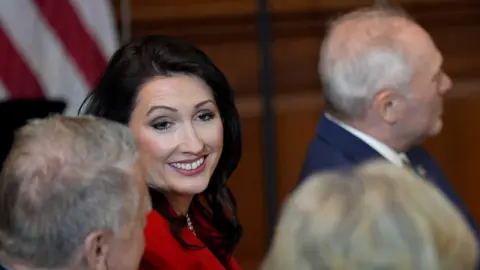 PA Media Emma Little-Pengelly. She has long dark hair, wearing a red blazer. Other people are sitting beside her.