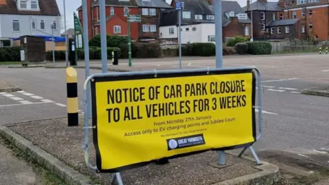 Great Yarmouth Borough Council A yellow sign is cable tied onto a piece of temporary fencing, stating: "notice of car park closure to all vehicles for three weeks" with smaller text giving details of dates and electric charging points. In the backdrop are houses and hotels.