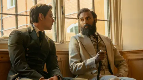 Netflix Louis Partridge and Himesh Patel in Enola Holmes sitting down talking to each other