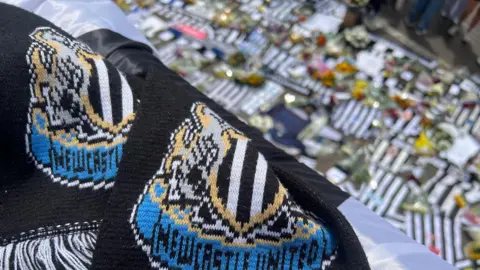 A Newcastle United scarf in the foreground. The background is blurred and shows tributes left at St James' Park for Kevin Keegan. They include team shirts and flowers.