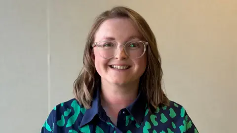 Alison Beattie looks straight at the camera. She is standing in front of a blank wall. She has shoulder-length brown hair and round glasses. She is wearing a green and blue patterned shirt