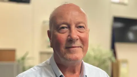 BBC Man with grey hair in shirt, standing in office.