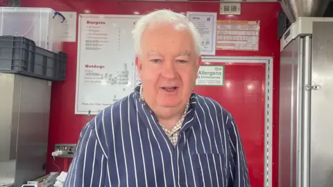 A man with grey hair and a dark blue shirt with white stripes in front of a red and silver background