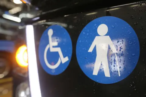 There are two blue badges on the side of a bus. One shows the image of a stick man with a walking stick. The other is the person in a wheel chair.