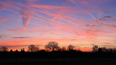 There are several trees and chimney tops silhouetted against the morning sky, which is turning blue but there is also a warm orange glow just above the horizon. Wisps of cloud shooting through the sky have also turned orange in colour.