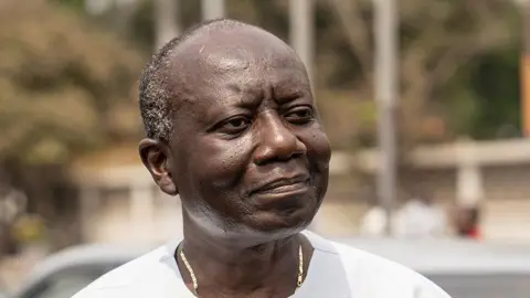 A head and shoulders image of Ken Ofori-Atta. He is wearing a white collarless shirt with a gold necklace just visible. He is looking to the right of the picture.