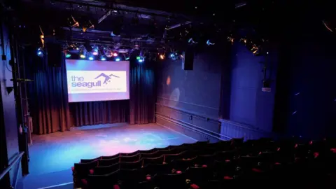 The Seagull A view of the Seagull Theatre from above the seats. A large stage can be seen as with a large screen at the back of it. 