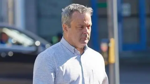 Christopher Elias walking in the sun to the court, he is wearing a white and grey checked shirt, has grey hair and has a furrowed brow.