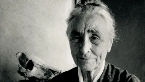 Black and white portrait of American artist Georgia O'Keeffe at her home in New Mexico, 1971. She has tied back hair and is looking past the person taking the photograph. 