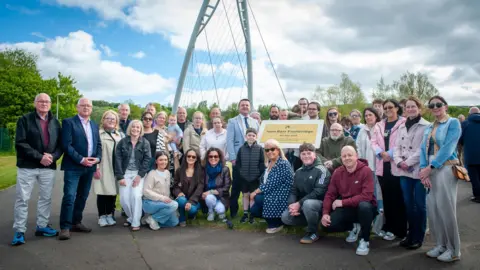 DCSDC A number of people, some standing and others kneeling down, are seen standing near to the golden plaque and the bridge is visible in the background.
