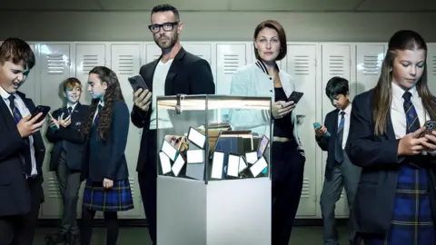 Channel 4 Matt and Emma Willis stand in a corridor holding smartphones. They are surrounding by young pupils wearing school uniforms. In front of the Willis' is a glass box with a number of smartphones in it. 