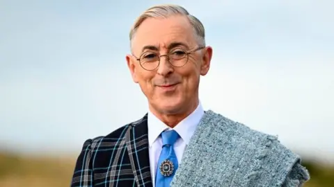 Alan Cumming smiling while wearing a blue tartan suit