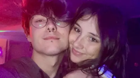 River Davies A young man with floppy dark hair, round glasses and a nose ring, with a young woman with long dark hair. Their faces are close together with their cheeks touching. Both are smiling and looking at the camera. The photo was taken in a club or pub at night. 