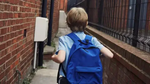 A boy walks down an alleyway by a building in a blue short sleeve shirt and blue back pack on.