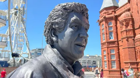 BBC A statue of Rhodri Morgan made of grey metal, his face looking upward slightly with his mouth open as though he is about to speak. You can also see the collar of an overcoat. The red Pierhead building and the ferris wheel attraction at Mermaid Quay are in the background with blue skies above. 