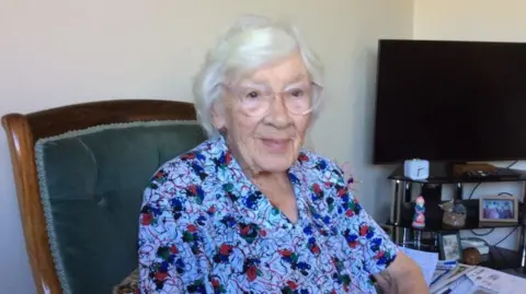 A white-haired woman wearing large plastic framed spectacles is seated in an upholstered armchair, slightly angled toward the camera. She is wearing a short-sleeved top with a floral pattern.