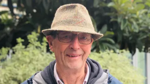 Peter Littlefield is standing outside in the sunshine. He is wearing a trilby hat, and glasses and is smiling at the camera. He is also wearing a white shirt, grey hoodie and blue anorak. 