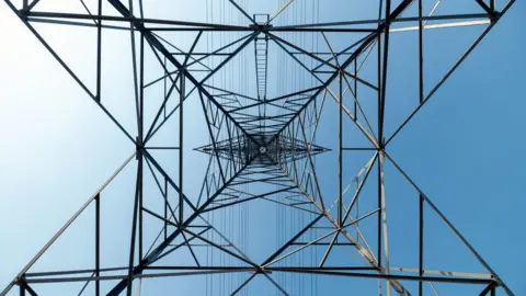 Library image of an electricity pylon. The camera is pointed upwards from the base of the structure. It is made of metal and has wires strung across horizontally. Blue sky can be seen in the background.