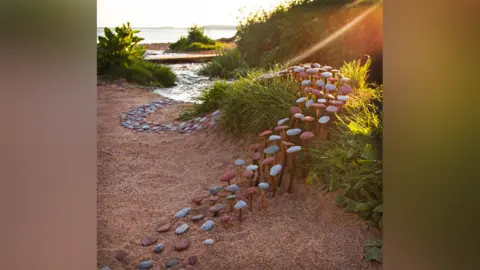 Jon Foreman/Sculpttheworld A pattern of pebbles that wind over sand and grass like a mini path, with water in the background and the glint of the sunshine. 