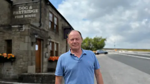 Steve Marsh is wearing a blue t-shirt and stood outside his pub the Dog and Partridge. There is a road, a car and trees in the distance.